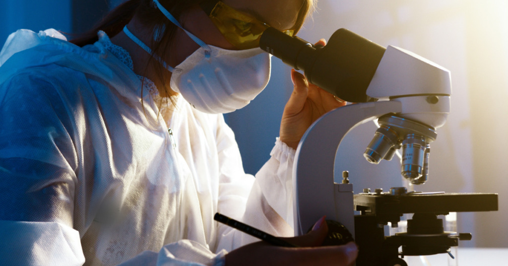 woman looking through a microscope with a mask and goggles on