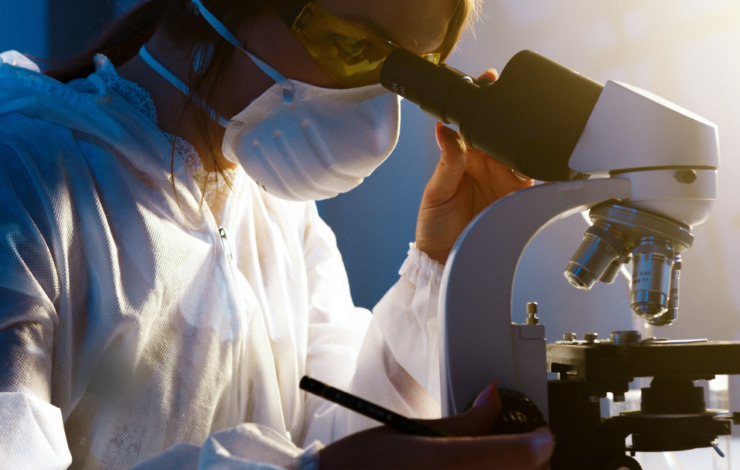 woman looking through a microscope with a mask and goggles on