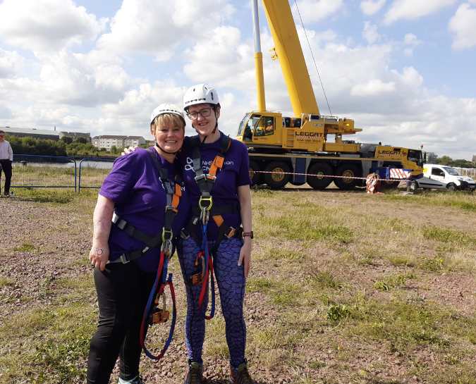 Two NHS Lothian Charity Champions stood near the beginning of the zipslide smiling at the camera. They have their harness and helmets on along with their NHS Lothian Charity purple t-shirt.