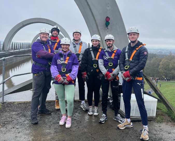 Seven NHS Lothian Charity Champions stood smiling at the camera waiting for their turn to do the abseil. Wearing a harness, helmet and a purple NHS Lothian Charity t-shirt.