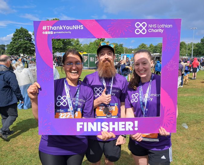 Three NHS Lothian Charity runners holding a finisher frame at the end of the EMF